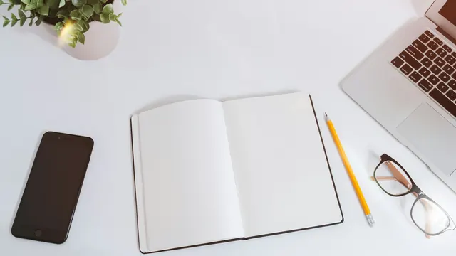 Aufgeschlagenes leeres Notizbuch, Smartphone, Bleistift, Brille und Laptop auf einem weißen Schreibtisch
