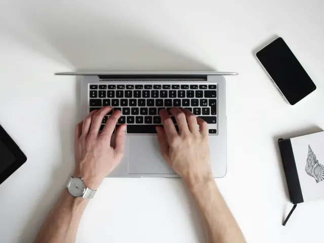 Dłonie piszące na klawiaturze laptopa na białym biurku, obok smartfon i notes